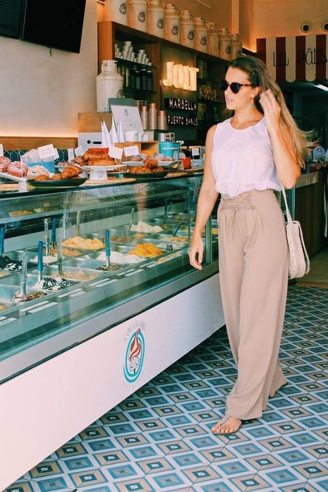 Woman in a gelato shop, looking at the display. Wearing sunglasses, top and pants, carrying a bag.