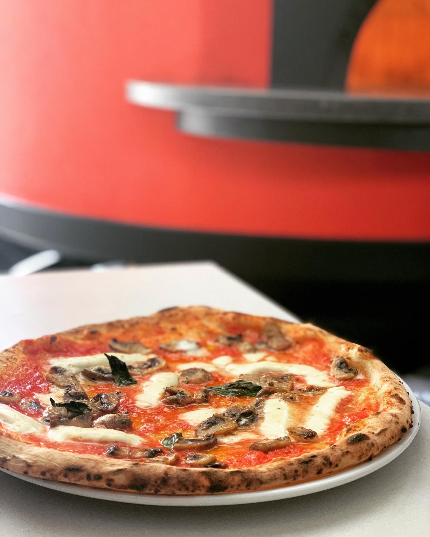 Pizza on a plate with mushrooms and cheese, in front of a red pizza oven.