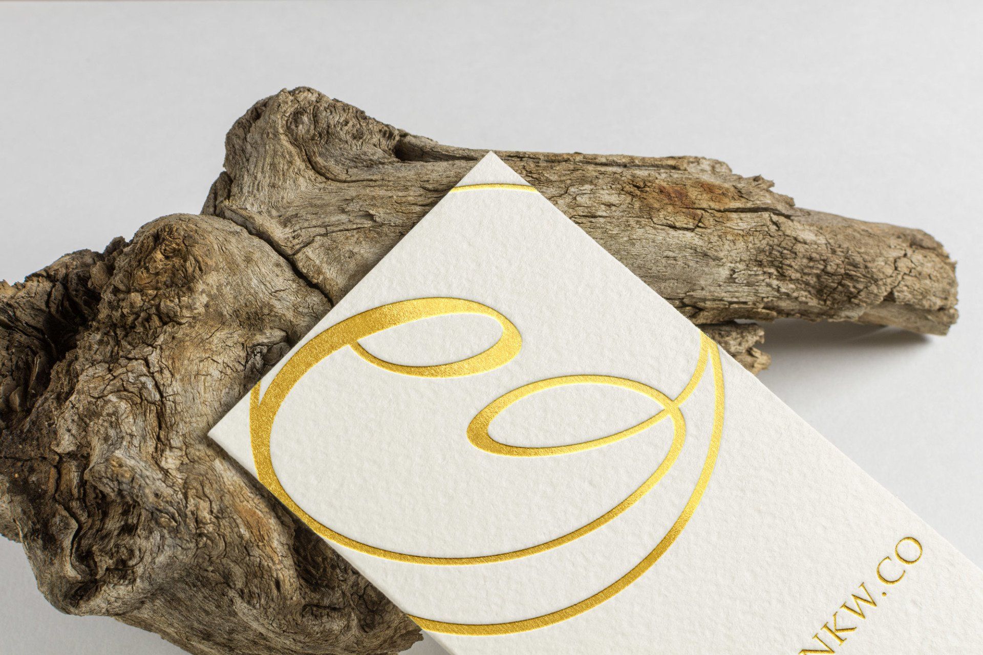 Cream business card with gold foil logo, resting on driftwood.