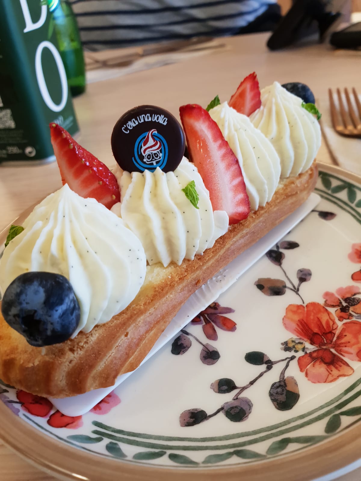 Eclair dessert with whipped cream, strawberries, blueberries, and a chocolate medallion on a floral plate.