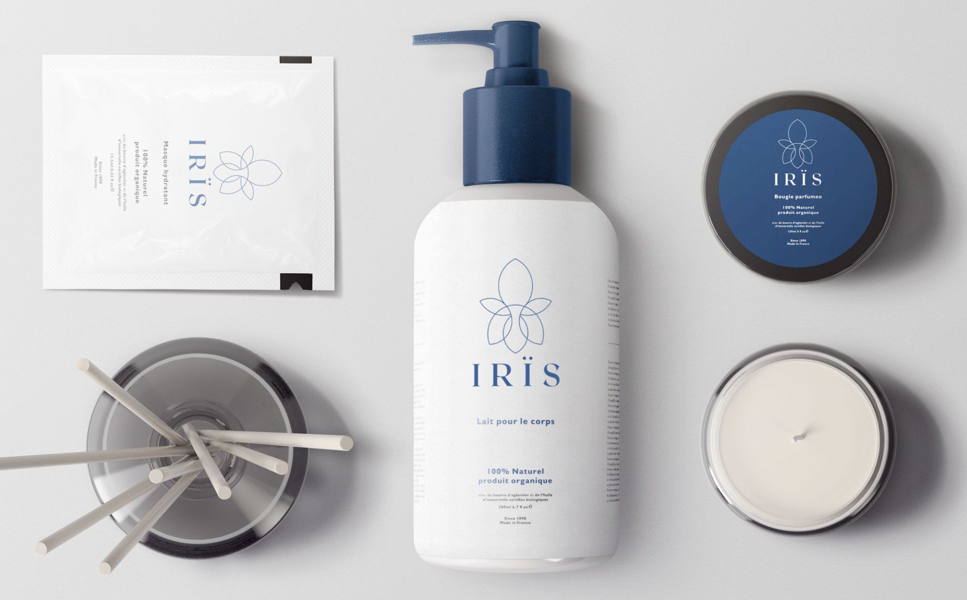 A flat lay of white Iris products: soap, candle, balm, and diffuser with blue accents on a grey surface.