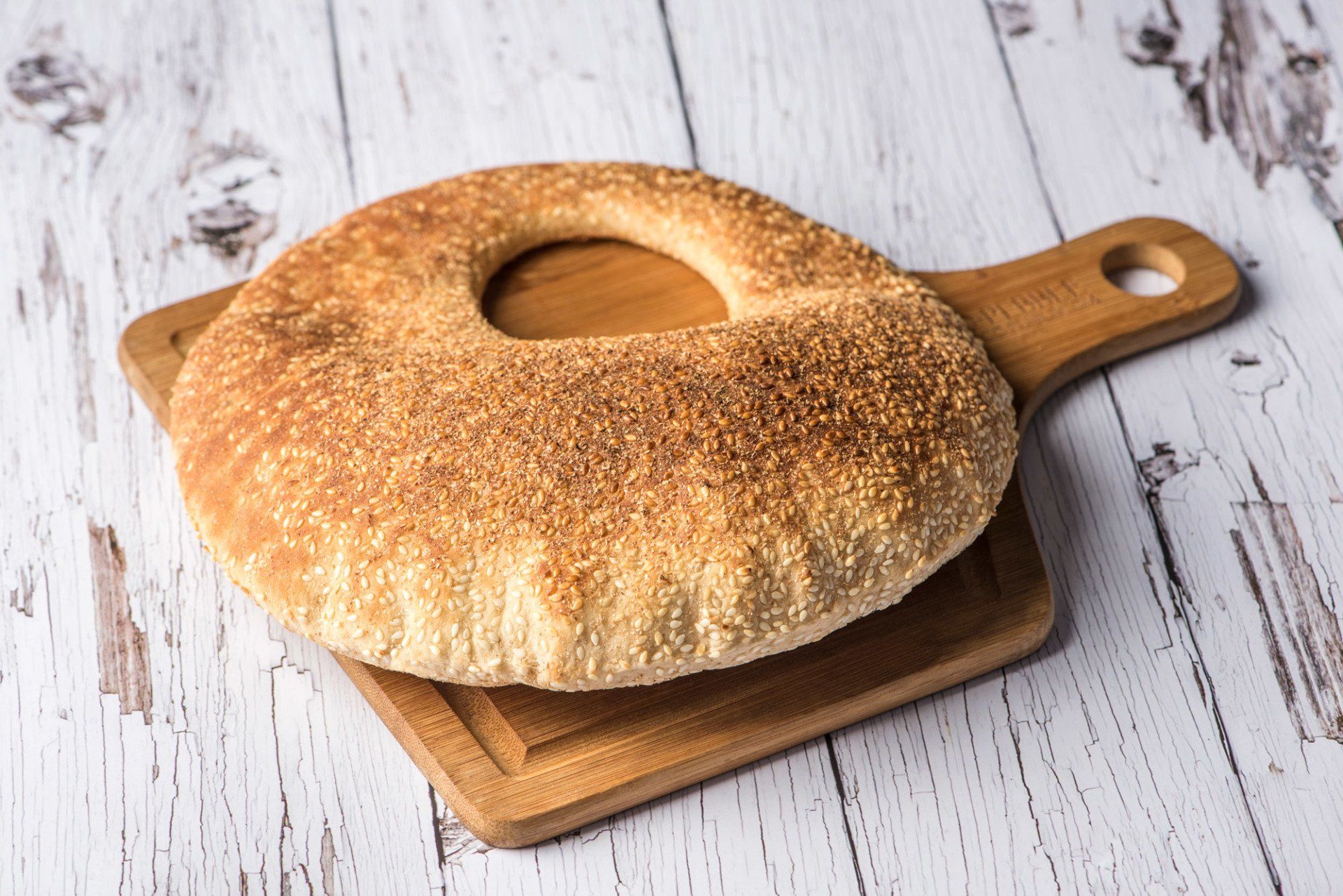 Round sesame bread with a hole on a wooden cutting board on a white wood surface.