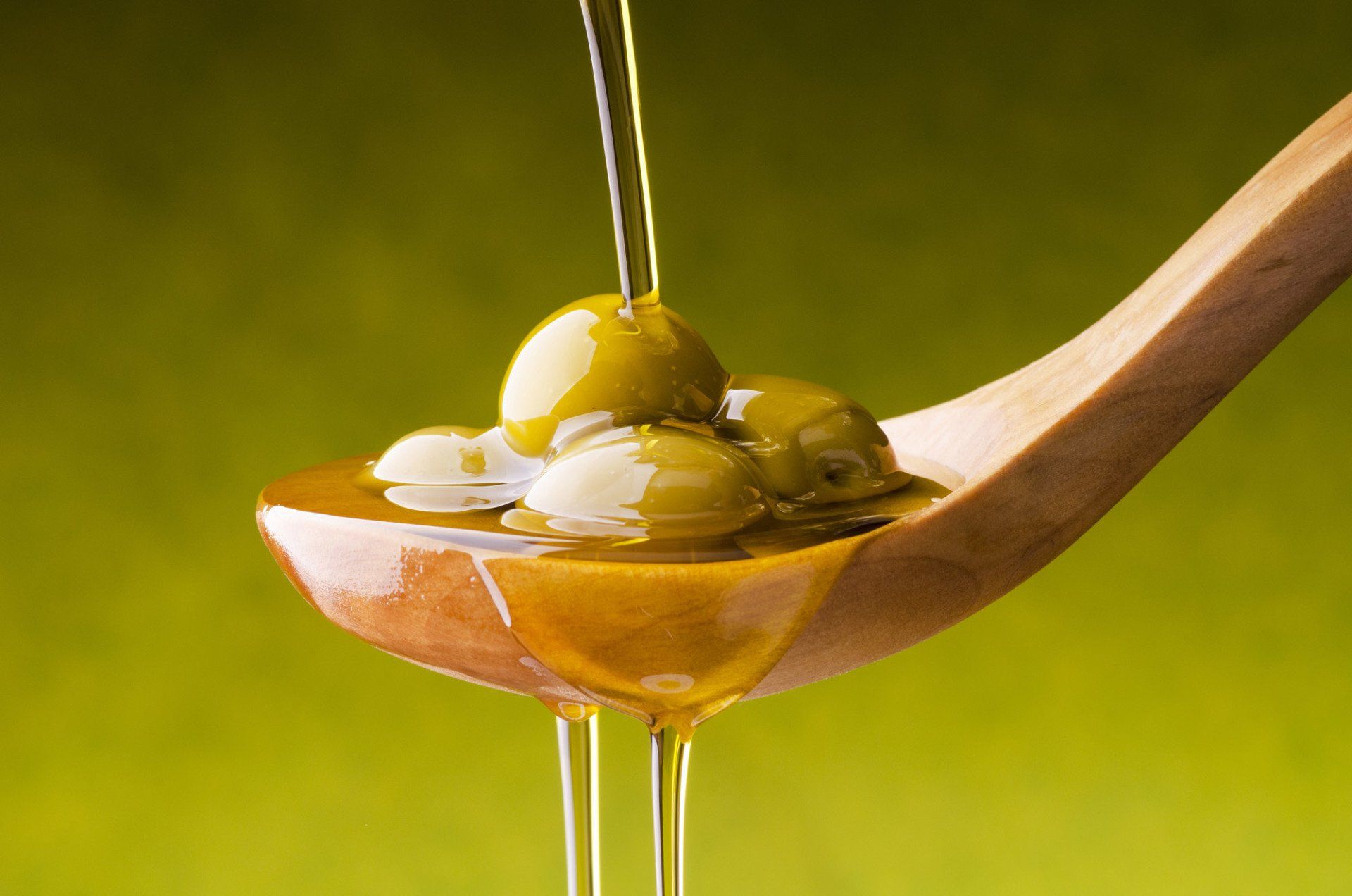 Green olives in a wooden spoon, olive oil being poured over them; green background.