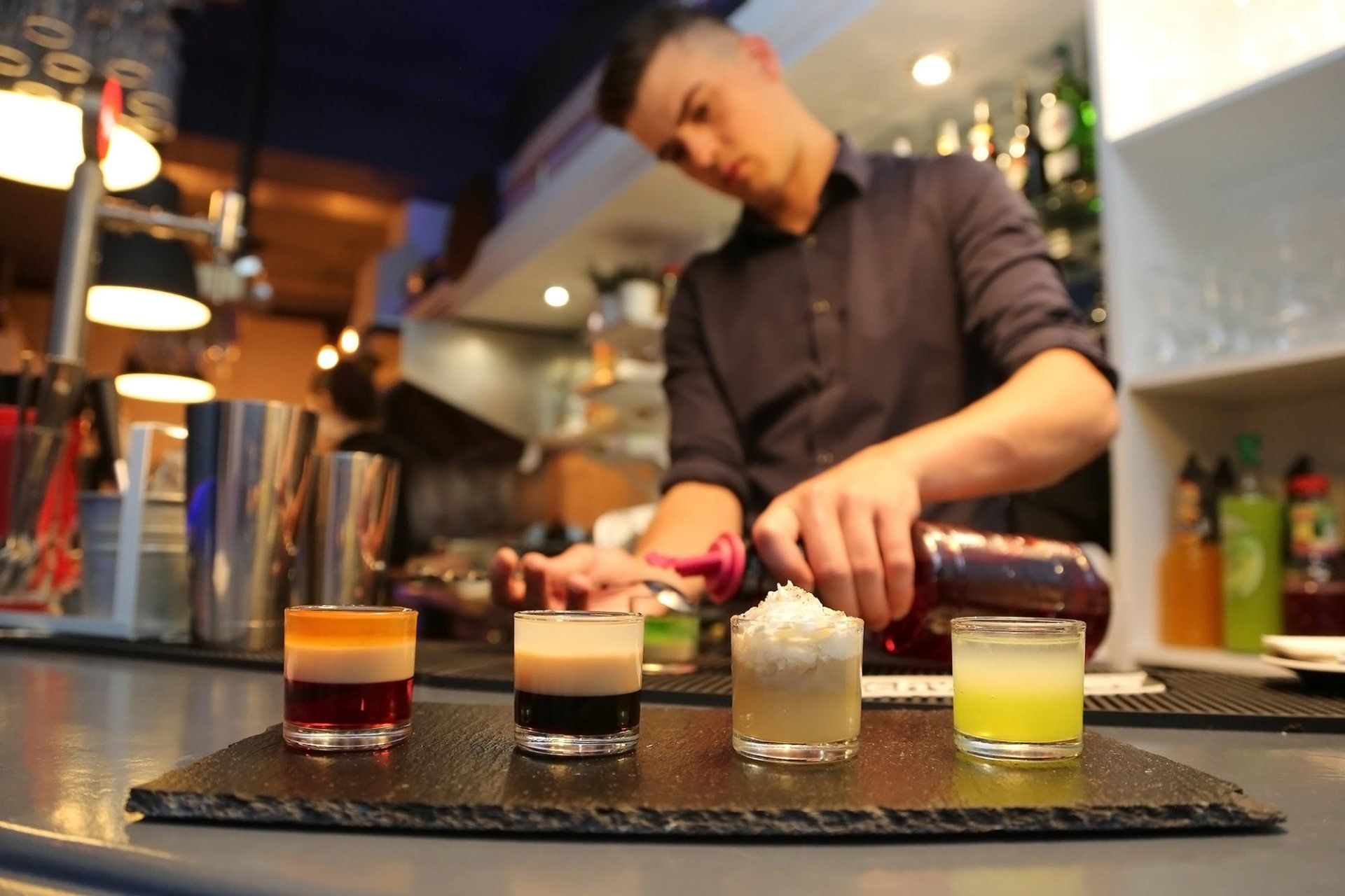Bartender pouring a drink while four colorful shots sit on a bar.