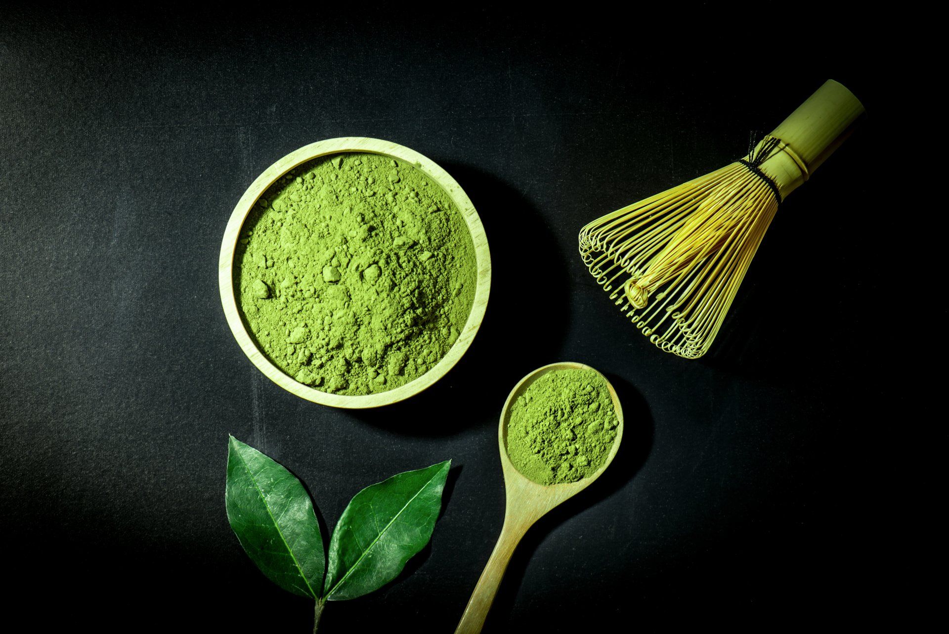 Matcha powder in a bowl and spoon, with a whisk, and leaves on a black surface.