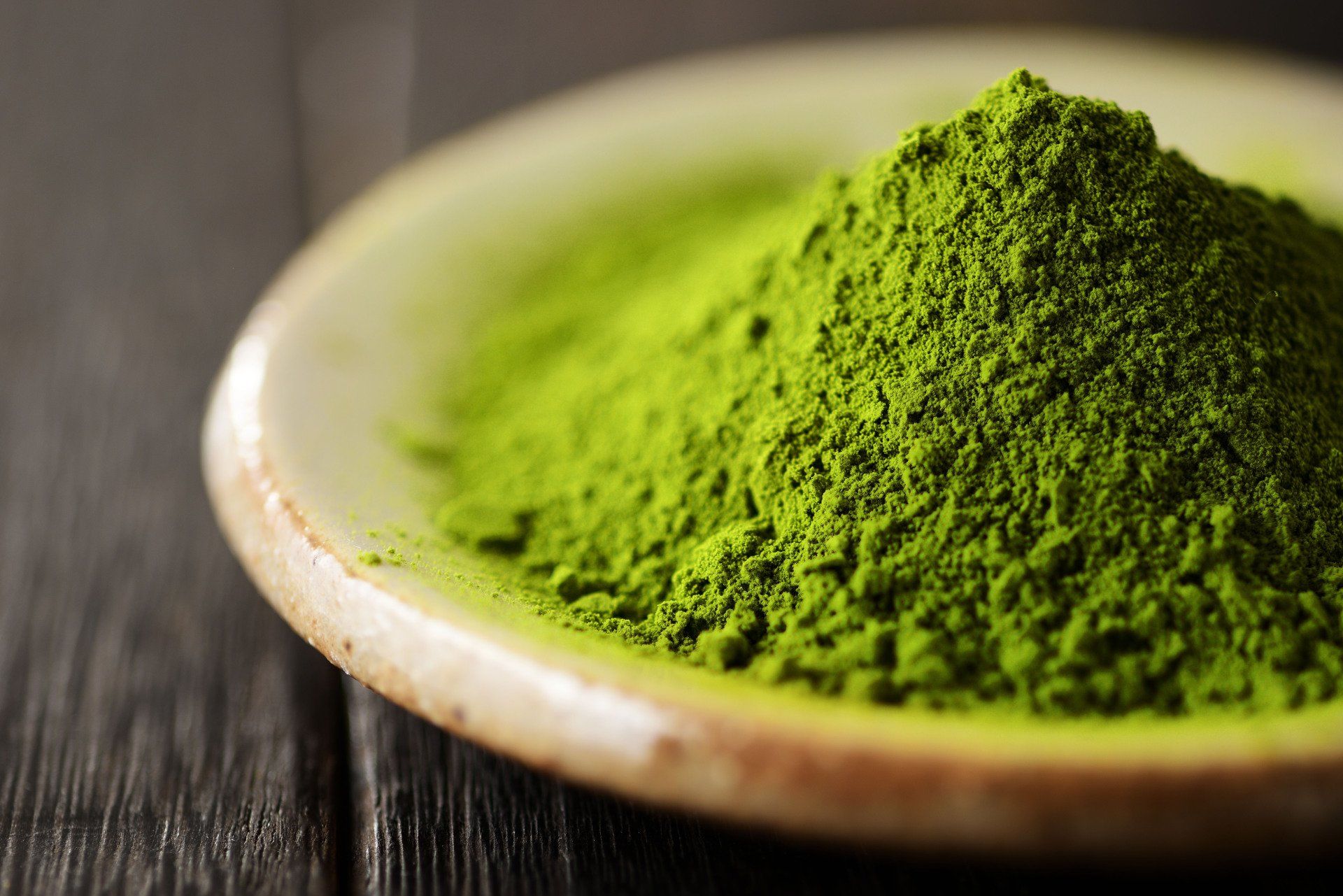 Green matcha powder piled in a small, off-white ceramic dish on a dark wooden surface.