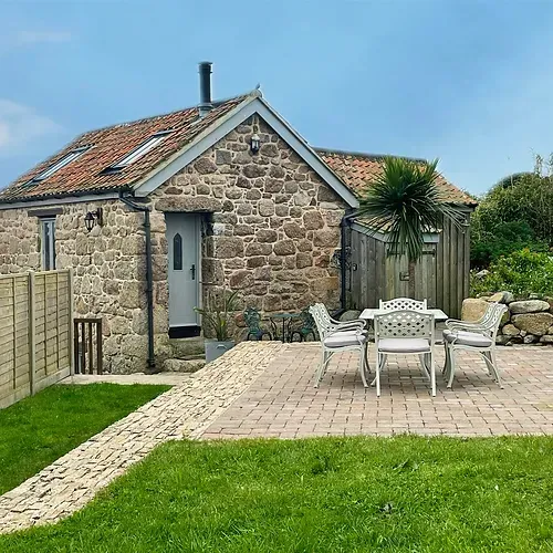 Stone cottage with patio furniture on a brick patio, surrounded by green grass and a wooden fence.