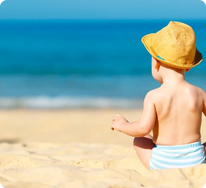Child wearing a yellow hat and blue-striped diaper sits on the beach, looking at the ocean.