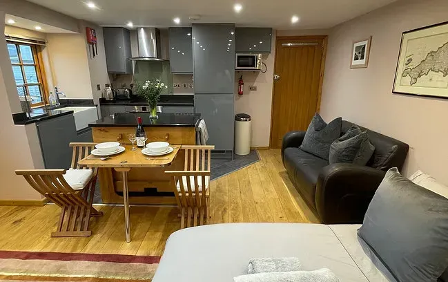 Studio apartment with kitchenette, seating, and bed. Dark gray cabinetry, wood floors, and a small dining set.