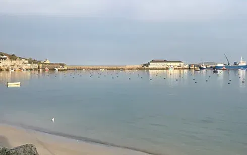 Calm harbor with a sandy beach, boats, and a distant building under a light blue sky.