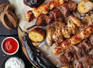 Grilled meats on skewers, eggplant, potatoes, and sauces on a wooden surface.