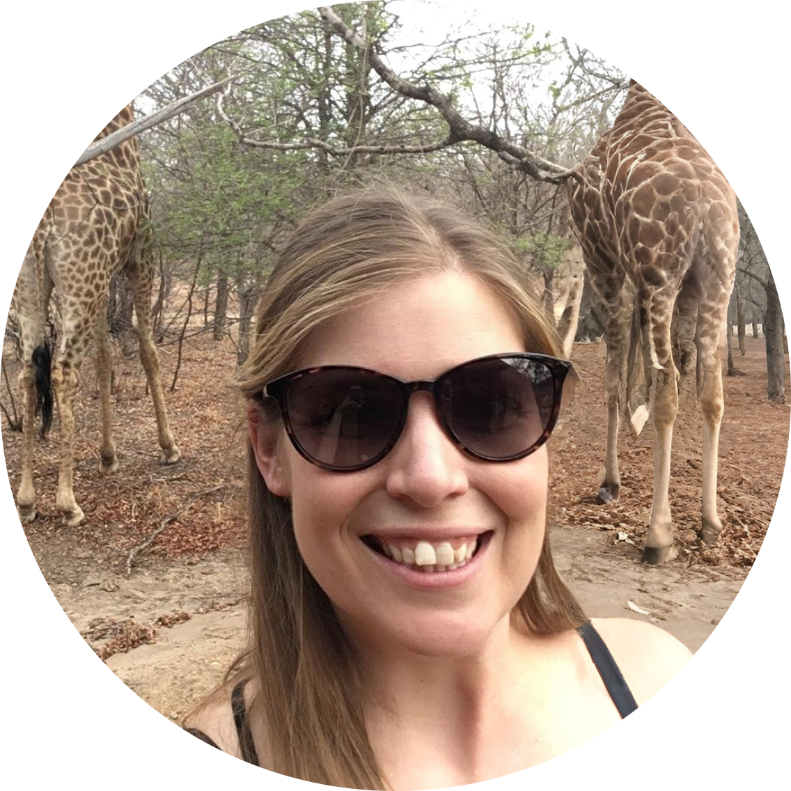 Woman smiling with giraffes in a natural setting, wearing sunglasses.