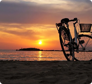 Bicycle on beach at sunset; orange and yellow sky, small island in water.