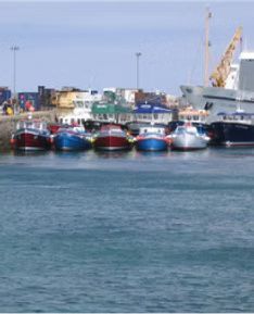 Boats docked at a harbor, with colorful buildings and a crane in the background under a blue sky.