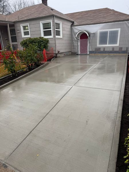 A newly poured concrete driveway leads to the front entrance of a grey, single-story house.