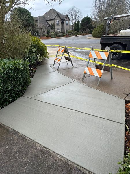 A freshly poured concrete sidewalk section is blocked off by two orange and white safety barricades.