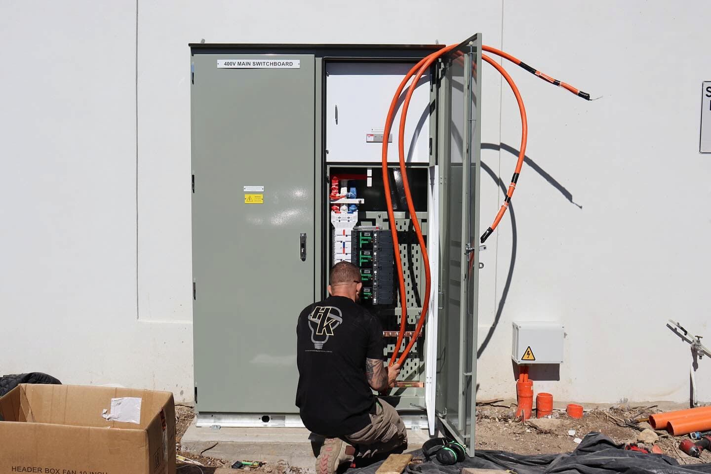 A worker kneeling to install thick orange cables into an outdoor electrical cabinet mounted on a white wall — Hspark Electrical PTY LTD in Wilton, NSW
