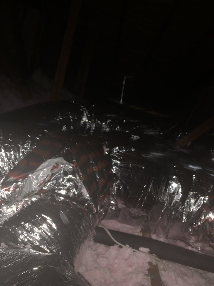 Flexible silver ductwork in a dark attic, resting on pink insulation, with wooden beams visible.