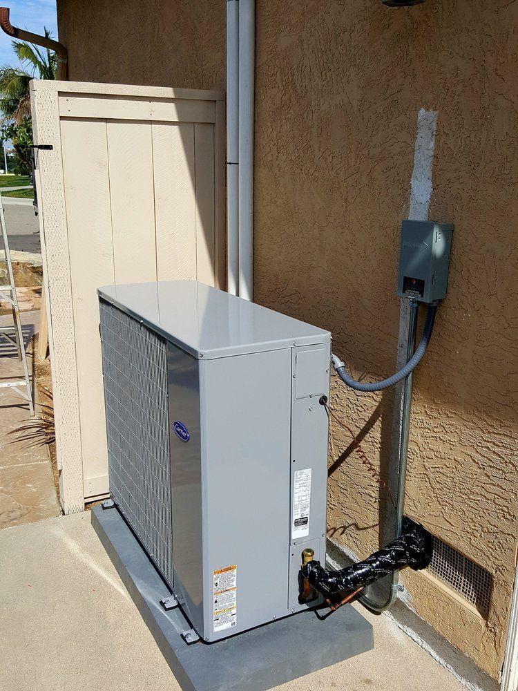 Gray air conditioning unit next to a light-colored wall and wooden fence, set on a concrete pad.
