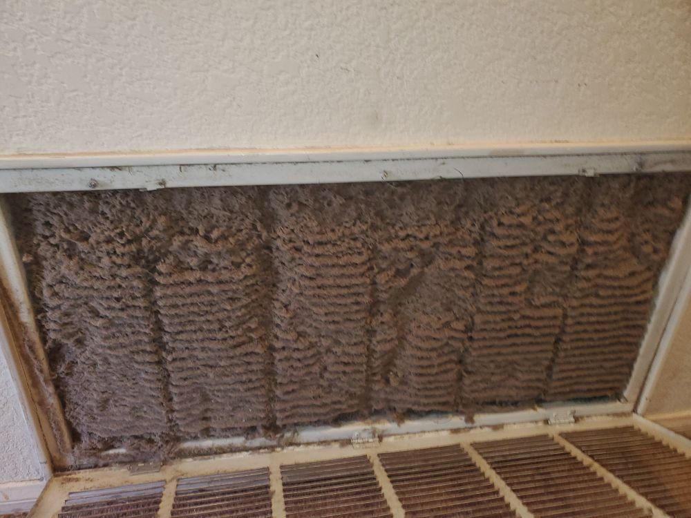 Dirty air filter in a white frame, clogged with thick, brown debris, set in a ceiling.