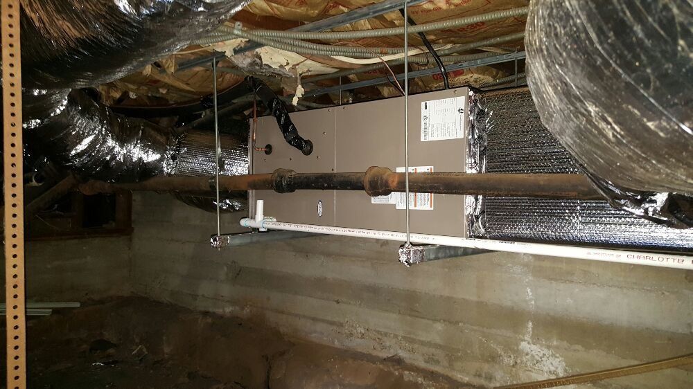 Air handler unit suspended in a crawl space, with ductwork and pipes visible.