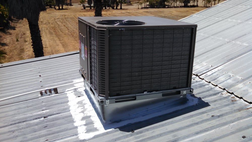 Air conditioning unit installed on a corrugated metal roof. The unit is gray. Outside in sunlight.