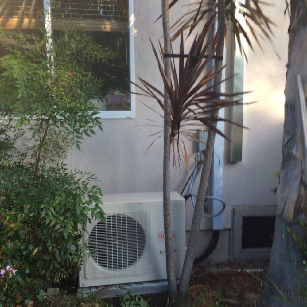 An air conditioning unit against a light-colored building, surrounded by plants and a window.