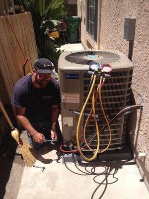 HVAC technician kneeling beside an air conditioner, using gauges, in an outdoor setting.