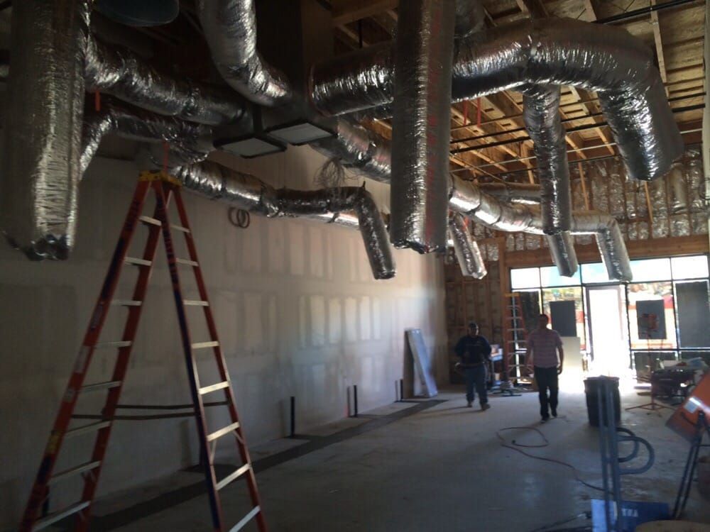 Interior of a building under construction, showing HVAC ducts, a ladder, and two people walking.