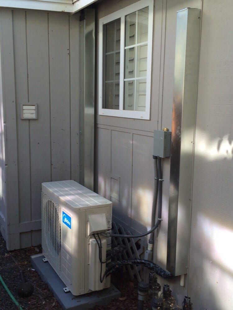 An air conditioning unit and metal exhaust pipes outside a building, near a window. Gray walls, overcast.