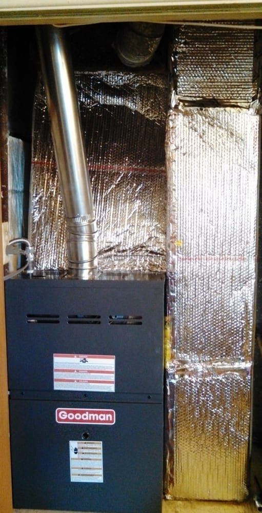 Furnace with a silver vent pipe in a utility closet with foil insulation. A Goodman brand furnace is visible.