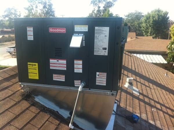 Goodman HVAC unit on a brown shingled roof with trees in the background.