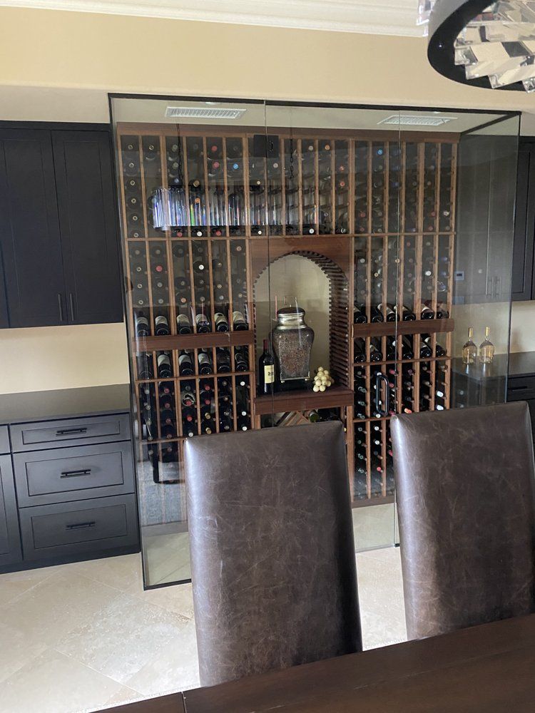 Wine rack with glass enclosure, brown bottles, wood, dark cabinets, two brown chairs in foreground.