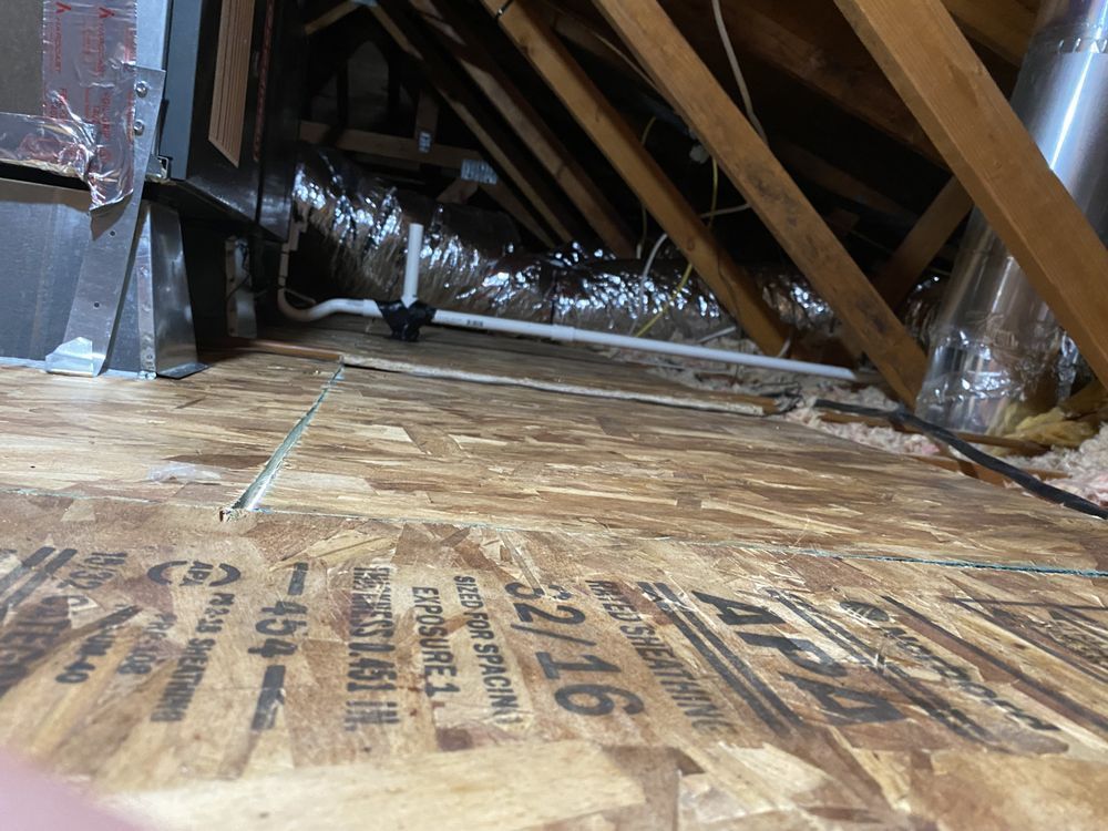 Inside of an attic with plywood flooring, rafters, HVAC components, and insulation visible.