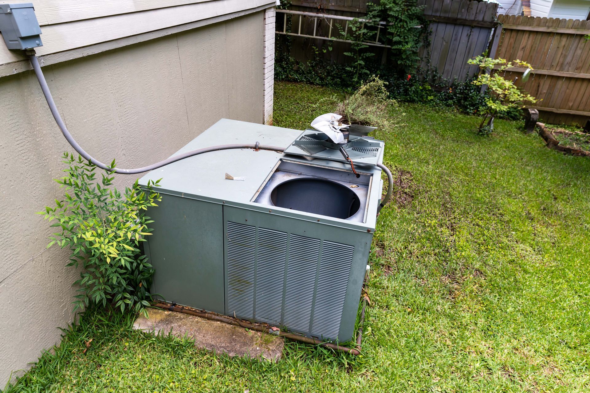 Air conditioning unit outside a beige building, surrounded by green grass and plants.