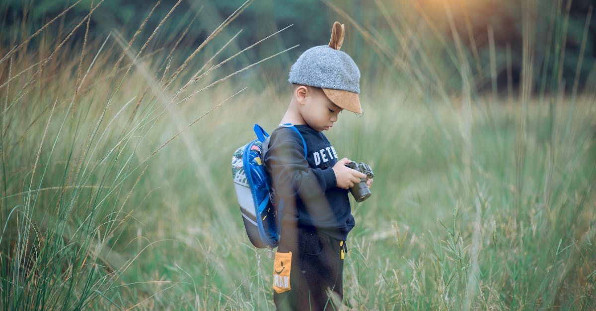 A young boy with a backpack is standing in a field holding a camera.