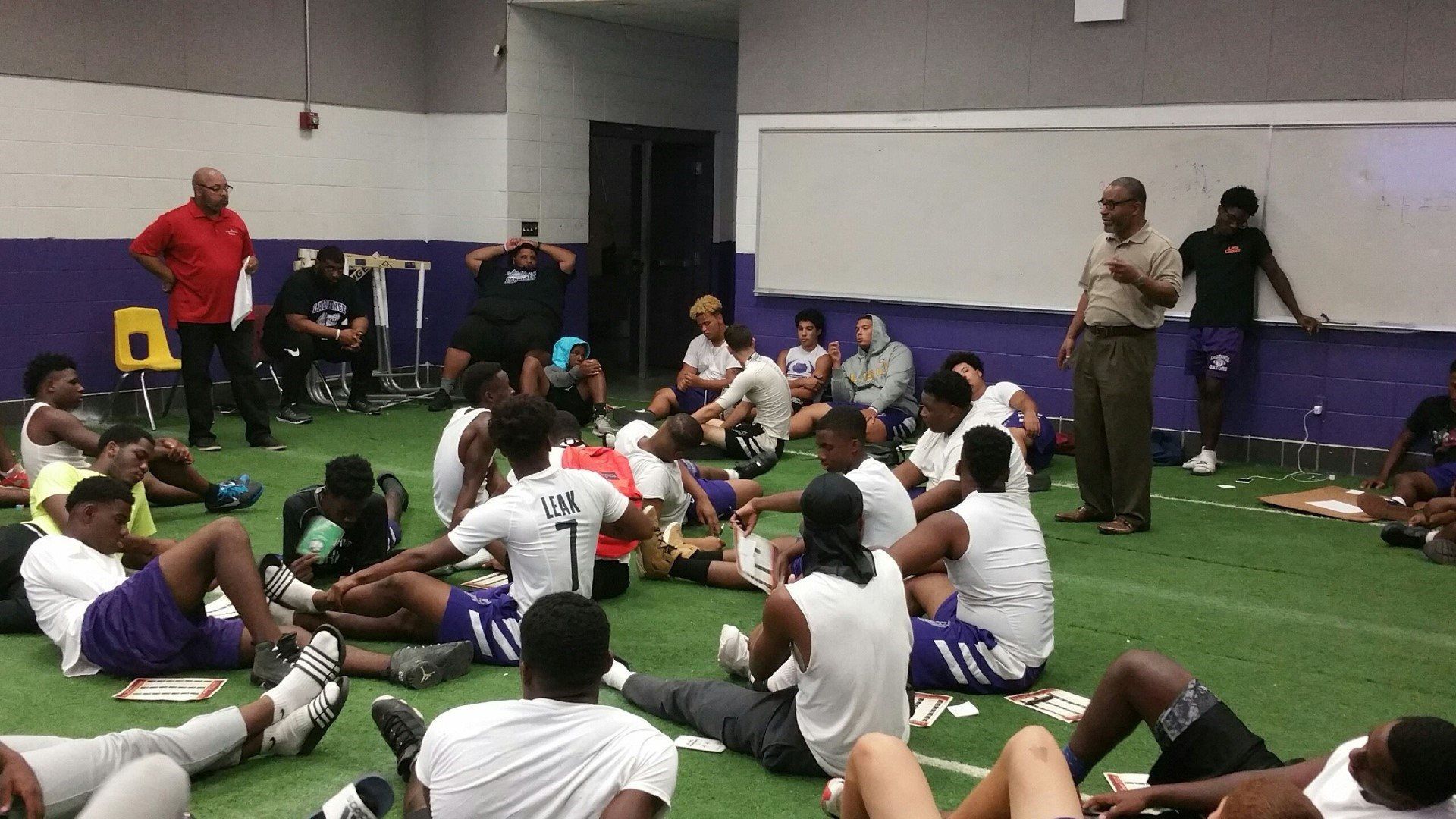 A group of young men are sitting on the floor in a gym.