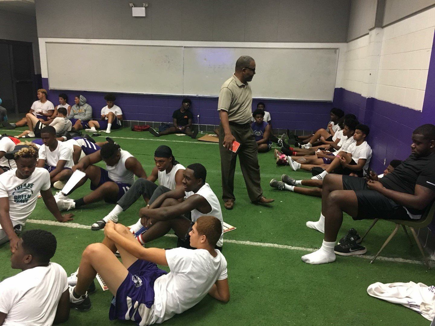 A group of people are sitting on the floor in a gym while a man stands in the background.