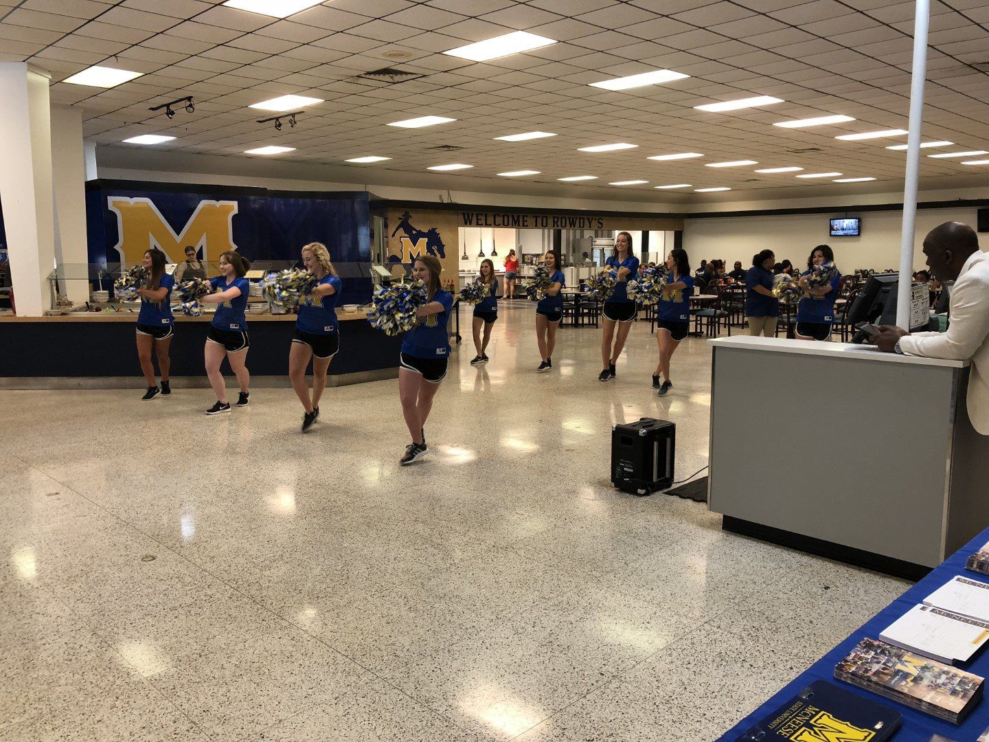 A group of cheerleaders are dancing in a room with a large m on the wall.