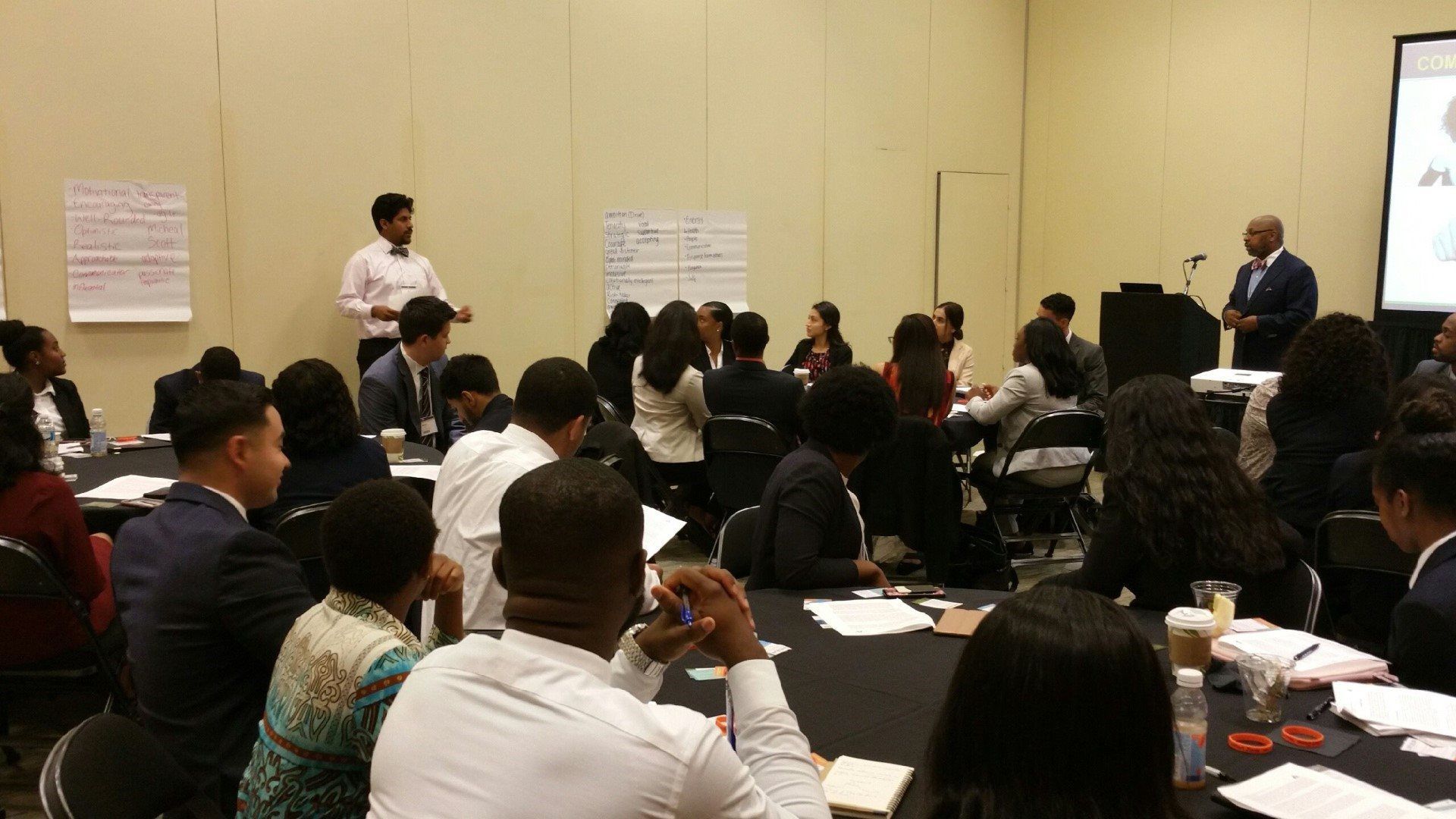 A group of people are sitting at tables in a room while a man gives a presentation.