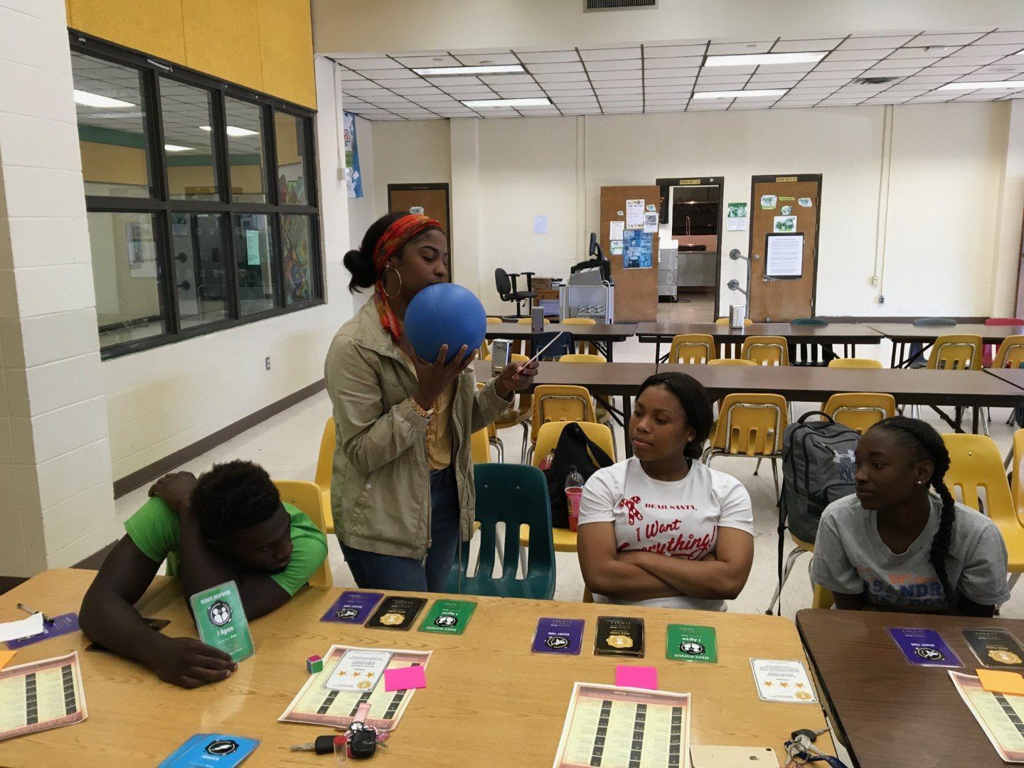 A group of people are sitting at a table with a woman blowing up a balloon.