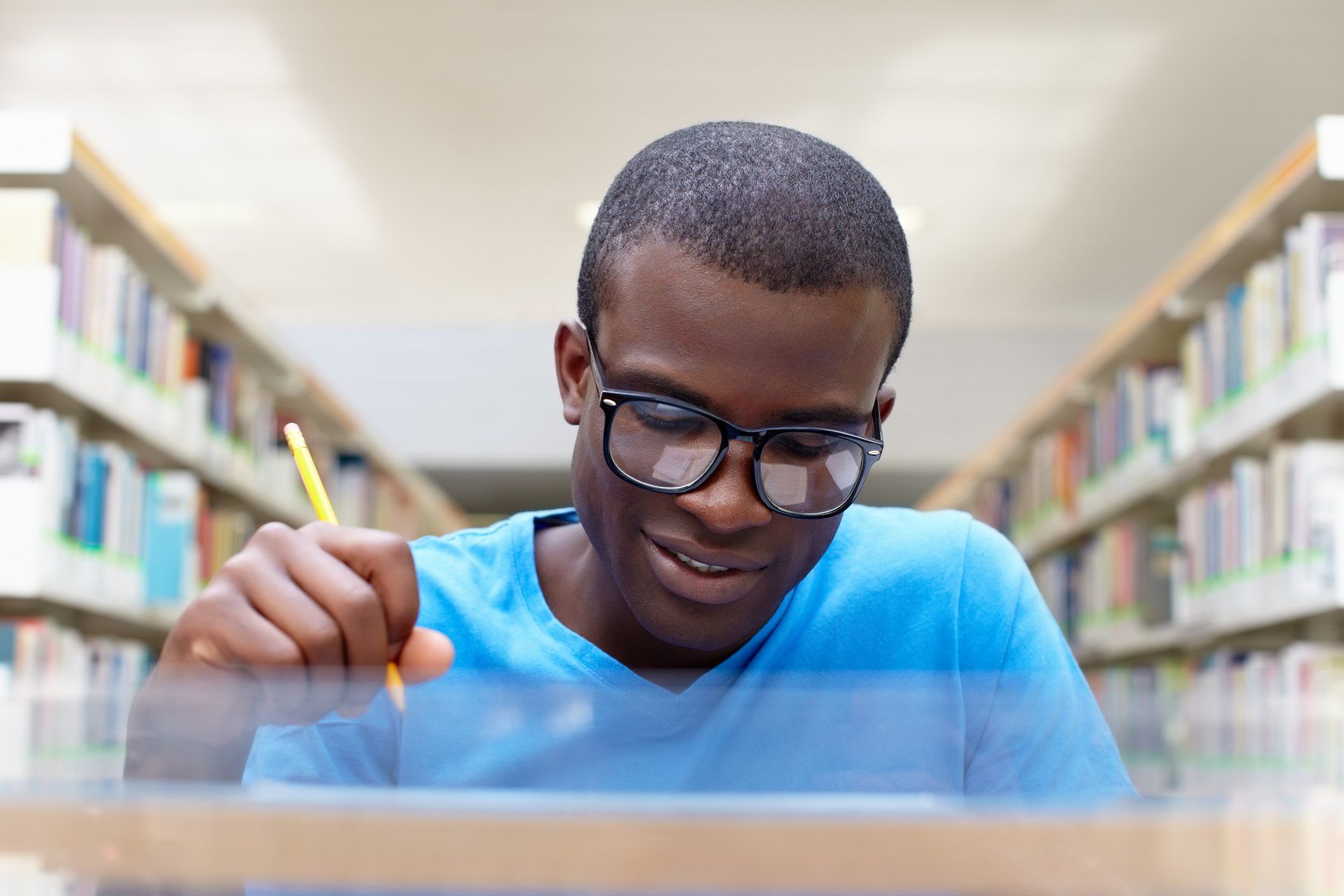 A young man is sitting at a desk in a library with a pencil in his hand.