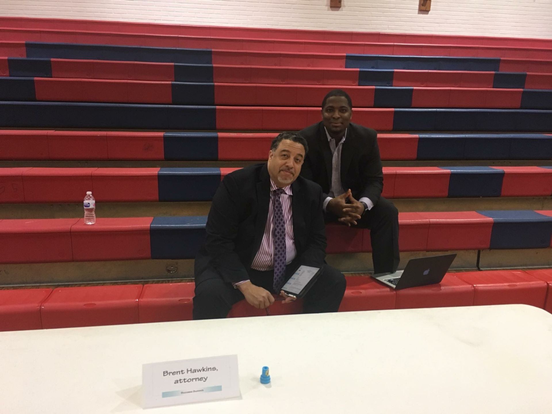 Two men are sitting on a bleacher with a laptop and a sign that says ' sam ' on it.