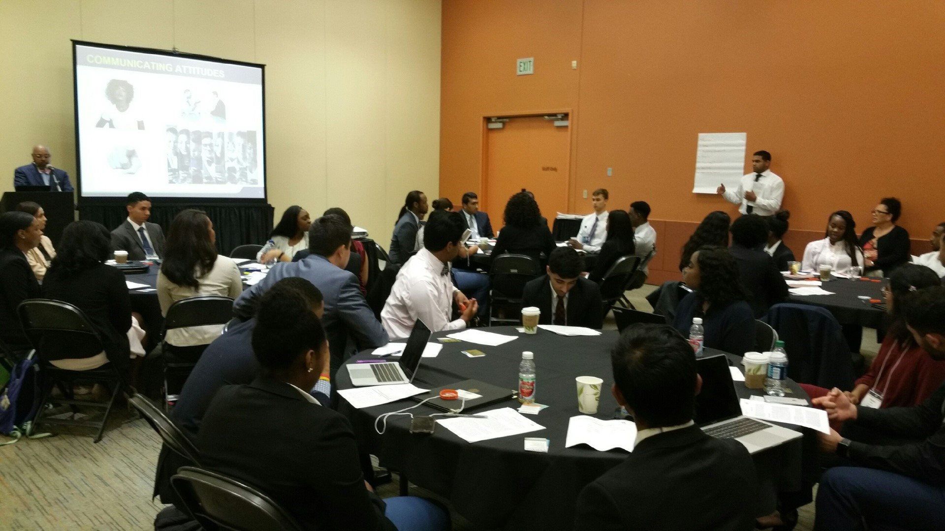 A group of people are sitting around tables in front of a projector screen