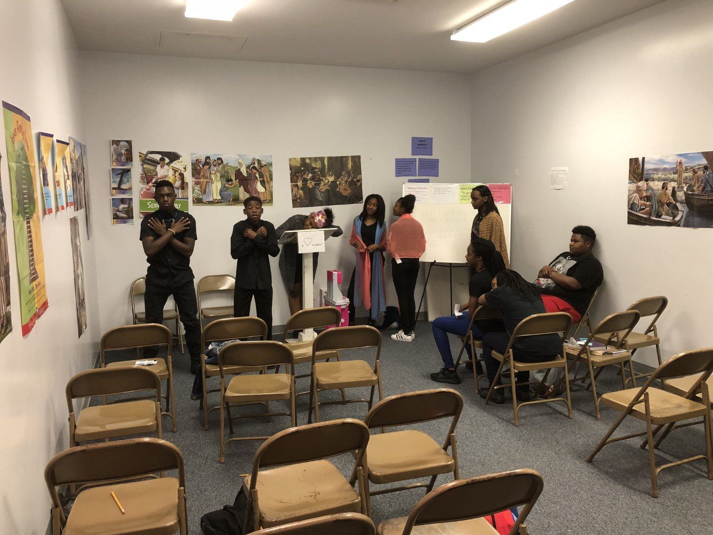 A group of people are standing and sitting in a room with folding chairs.