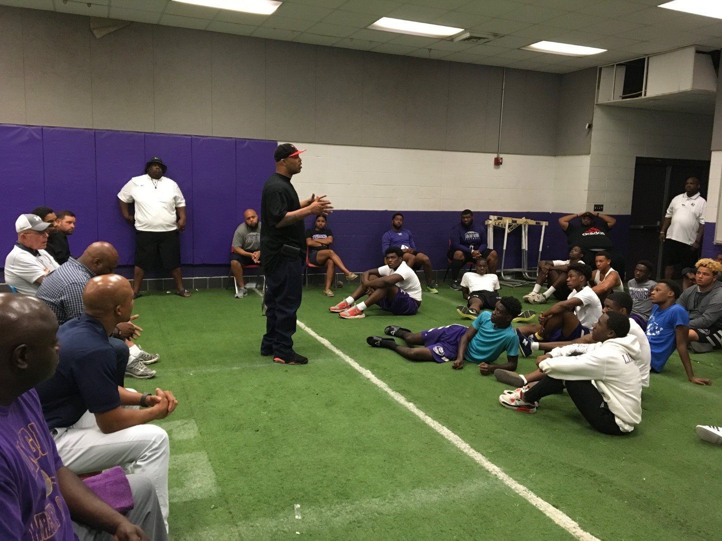 A man is talking to a group of people sitting on the floor in a gym.