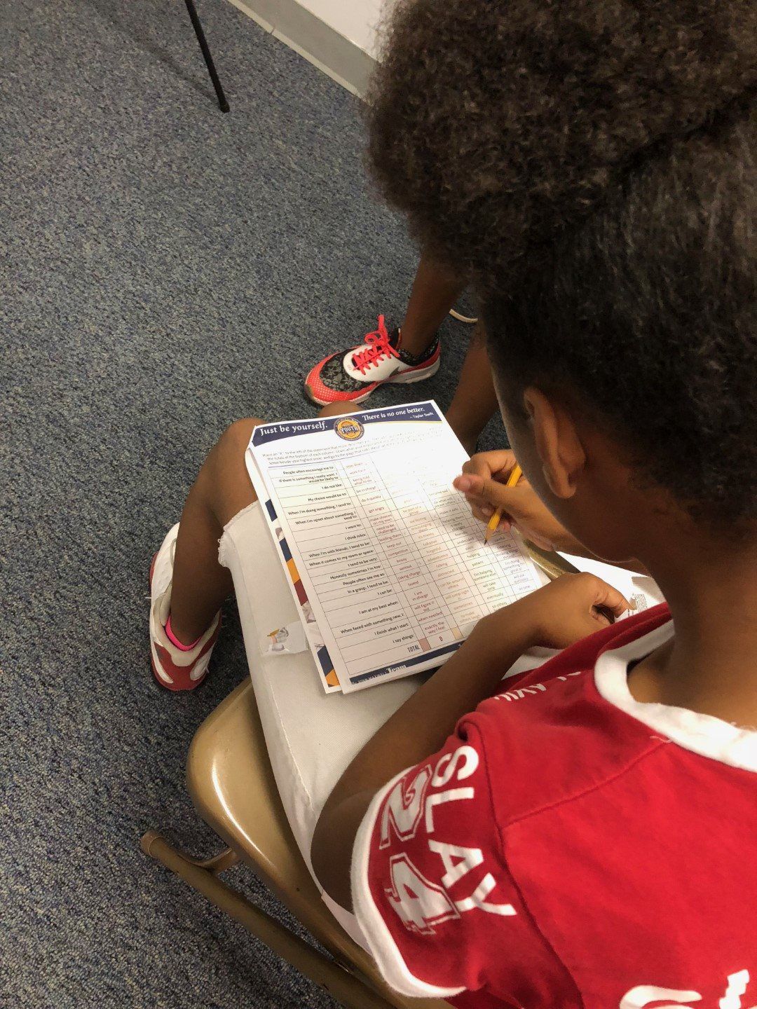 A girl in a red shirt is sitting in a chair writing on a clipboard.