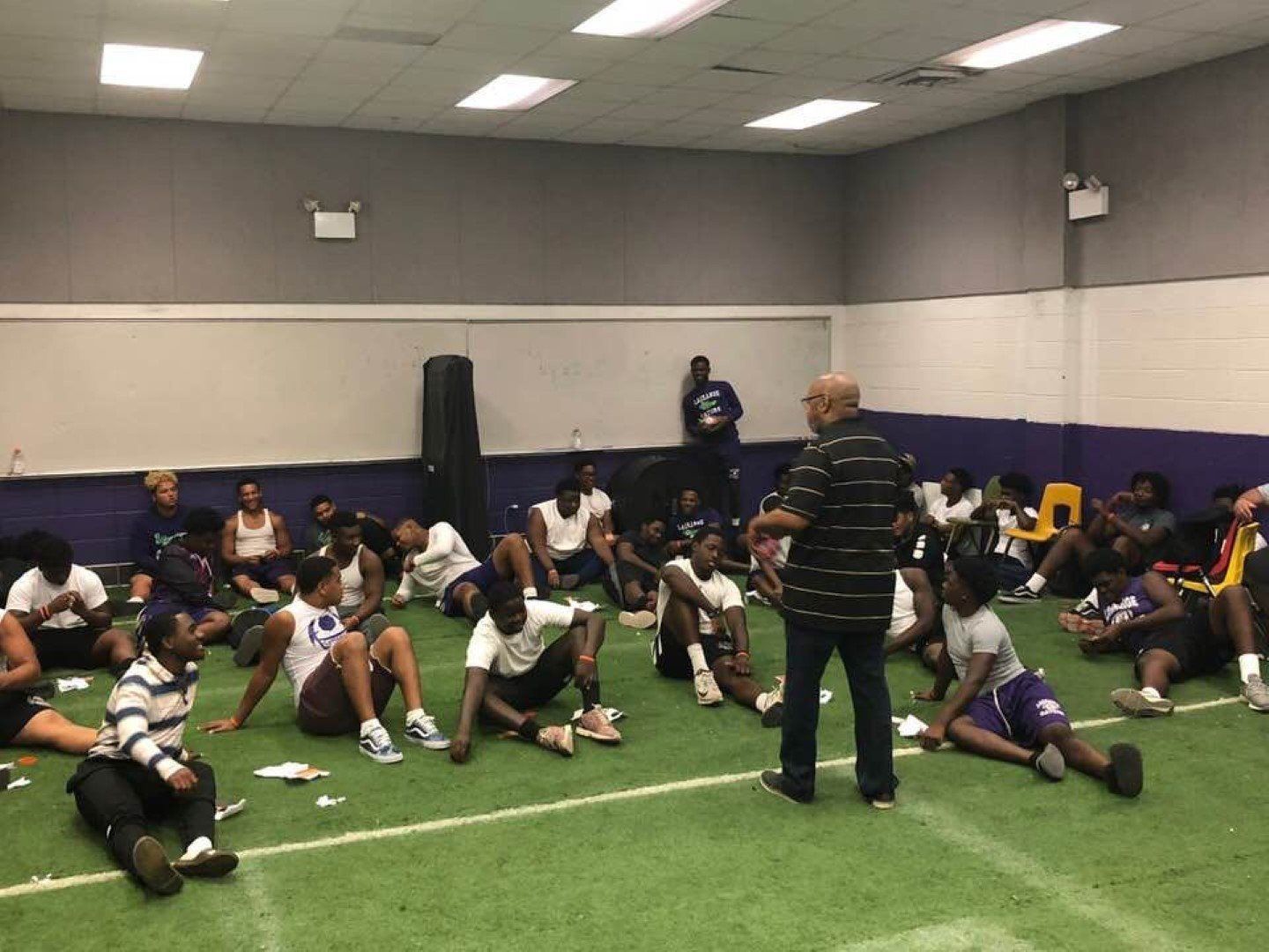 A man is talking to a group of people sitting on the floor in a gym.