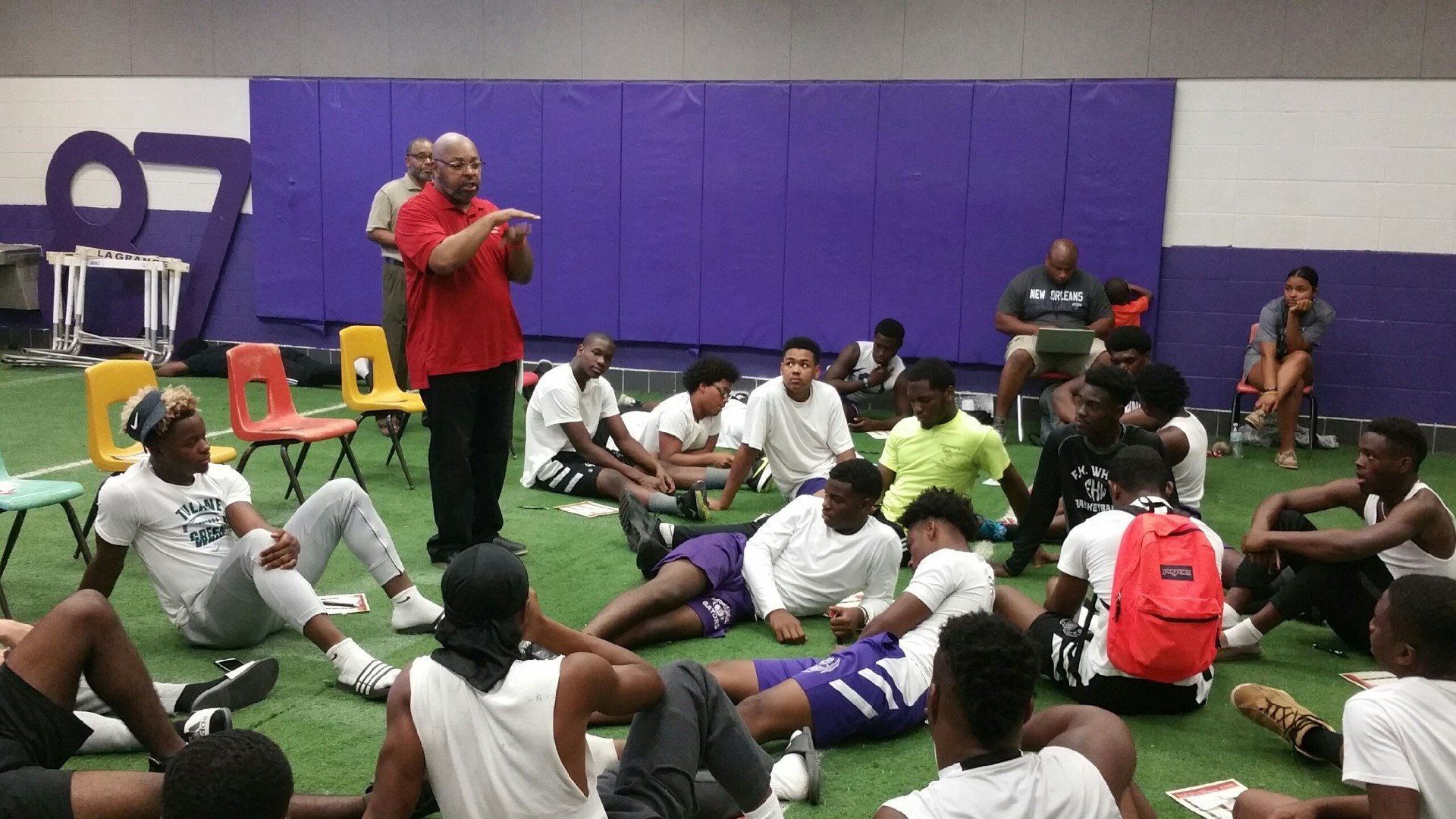 A man in a red shirt is talking to a group of young men sitting on the floor.