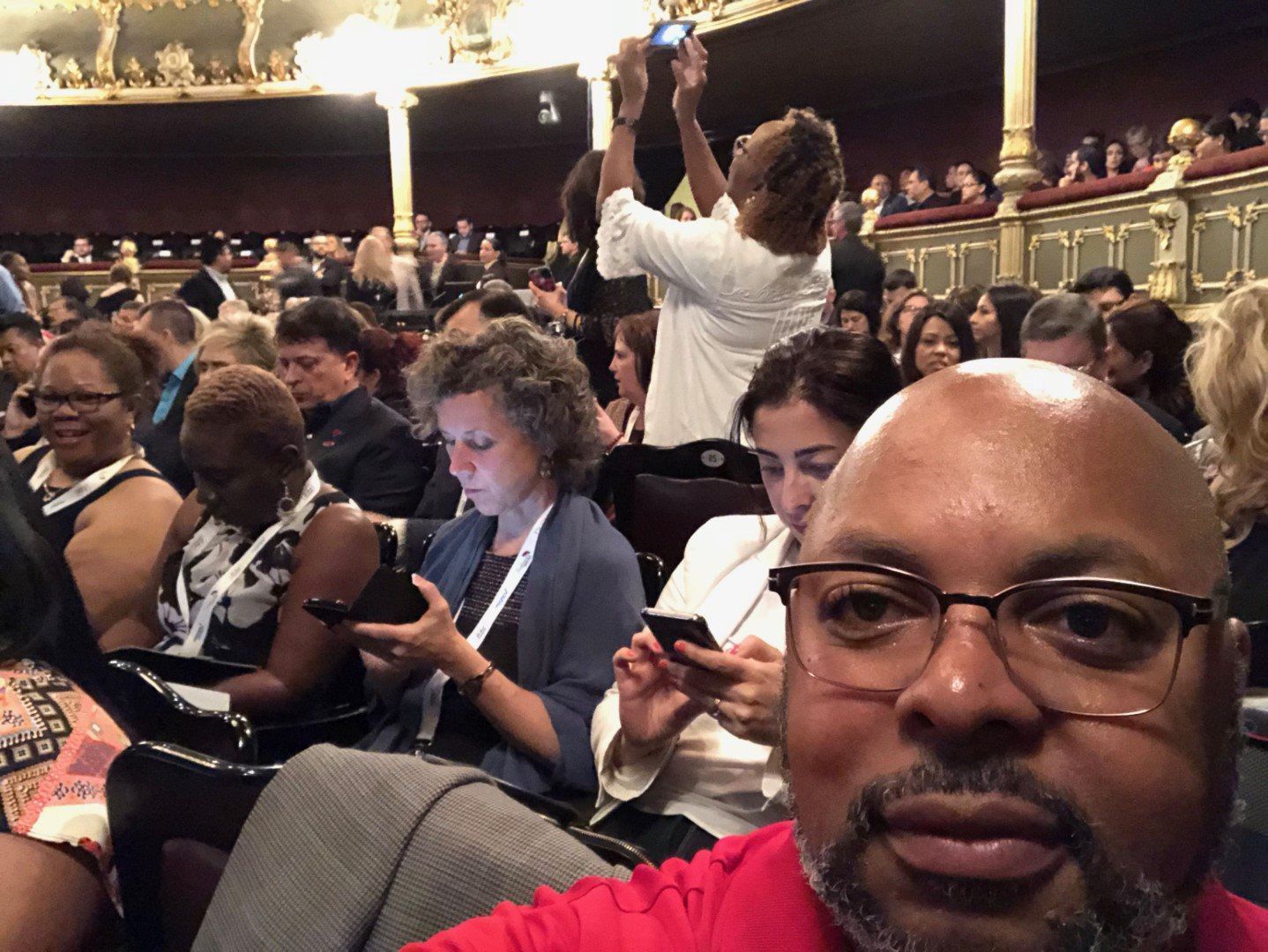 A man in a red shirt is taking a selfie in front of a crowd of people